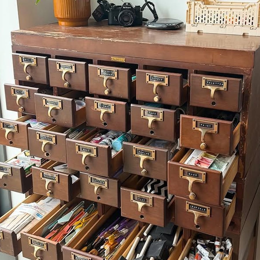 A vintage wooden filing cabinet with multiple open drawers, each filled with various arts and crafts supplies. A camera and a crate are sitting on top of the cabinet.