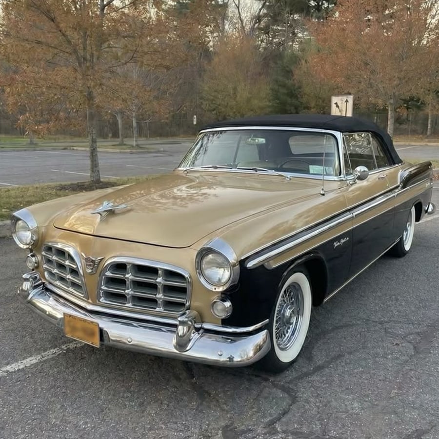 A vintage gold and black convertible car with whitewall tires is parked in an empty lot surrounded by autumn trees.