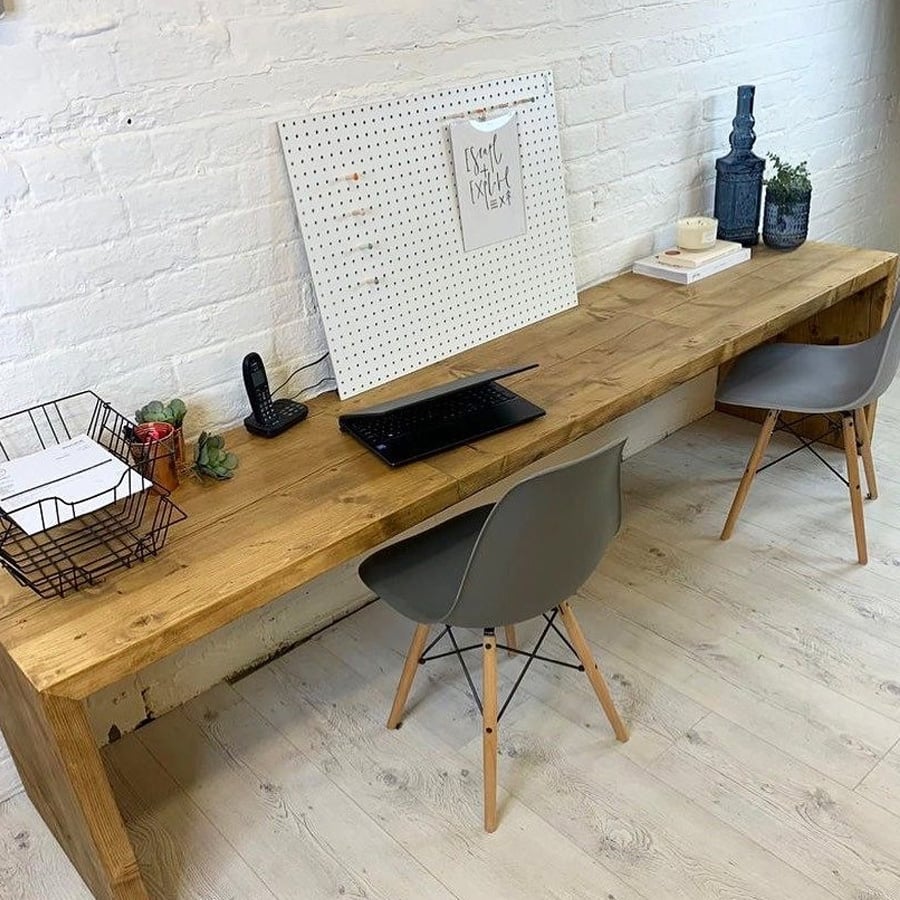 A wooden desk with two gray chairs, a wire basket with papers, a pegboard, a phone, a keyboard, plants, books, and decorative items, set against a white brick wall on a light wood floor.