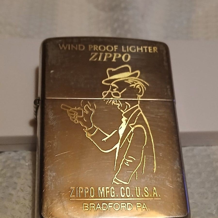 A gold Zippo lighter with an engraved image of a man in a hat pointing and text reading "WIND PROOF LIGHTER ZIPPO" and "ZIPPO MFG. CO. U.S.A. BRADFORD PA.