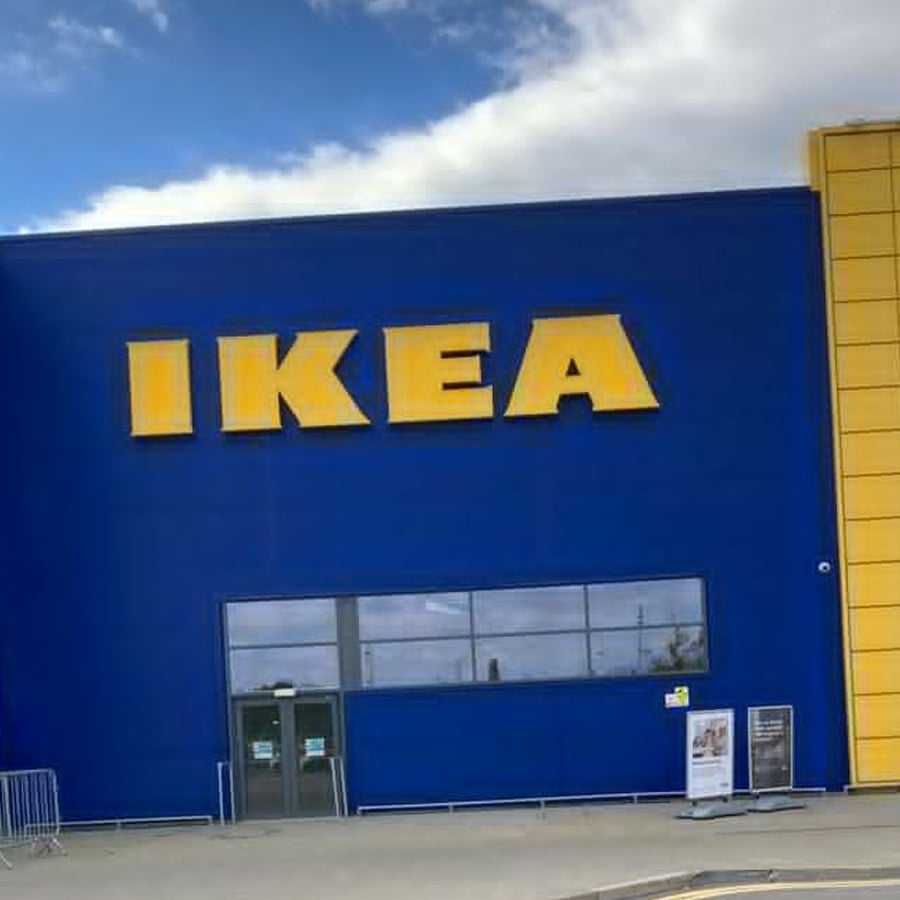 The exterior of an IKEA store with blue walls, large yellow "IKEA" letters, and a yellow accent on the side. There are windows, entrance doors, and a cloudy sky above.