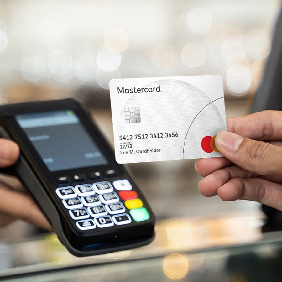A person holds a Mastercard credit card near a card reader, ready for a contactless payment. Another hand is visible next to the payment terminal. The card details and keypad are clearly shown.