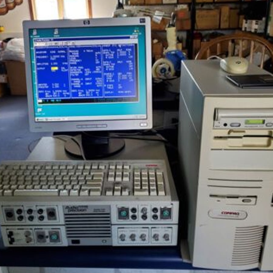 A vintage desktop computer setup with a CRT monitor displaying a blue interface, a beige Compaq tower, and an old keyboard with multiple dials and ports, all placed on a desk in a cluttered room.