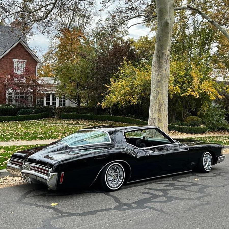 A shiny black vintage car is parked on a residential street lined with autumn trees, with a red brick house and green bushes in the background.