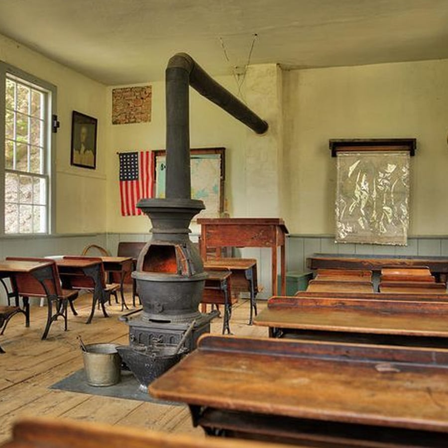 Old-fashioned classroom with wooden desks, a central wood stove, a U.S. flag on the wall, large windows, and a teacher’s desk at the front. Warm, vintage atmosphere with natural light coming in.