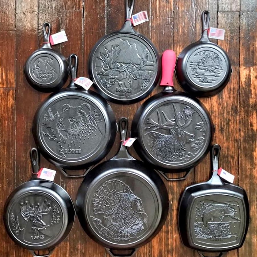 Eight cast iron pans with wildlife designs and small American flags on the handles are arranged on a wooden floor. Designs include animals such as bears, elk, and eagles.