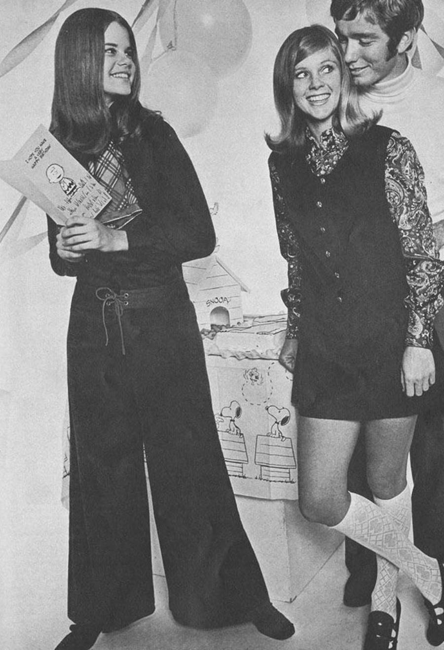 Three young adults at a party, two women and one man, all smiling. One woman wears a long-sleeve top with wide pants and holds gifts. The other wears a patterned blouse, a short dress, and knee-high socks. The man stands behind them, smiling.