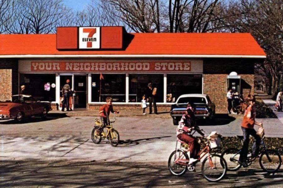 Children ride bikes past a 1970s-era 7-Eleven store with vintage cars parked outside. The building has a red roof and a sign reading "YOUR NEIGHBORHOOD STORE." Leafless trees fill the background.