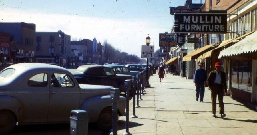 A vintage street scene shows parked classic cars, people walking on the sidewalk, and storefronts with signs, including one for Mullin Furniture, on a sunny day.