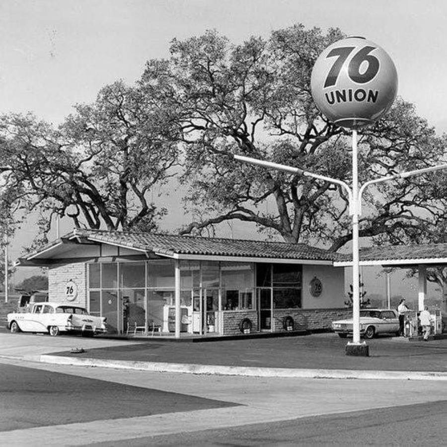 Black-and-white photo of a vintage Union 76 gas station with two classic cars parked outside, large trees in the background, and a round 76 sign on a tall pole in the foreground.