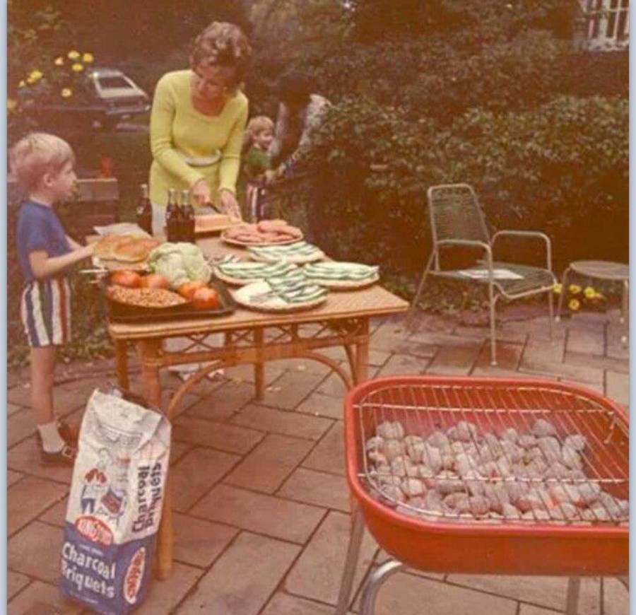 A woman and two children prepare food on a patio table near a grill with charcoal. Watermelon, lettuce, and soda bottles are on the table. Patio chairs and flowers are in the background.