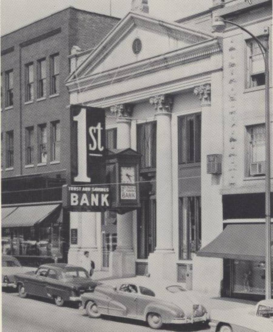 Black-and-white photo of a 1950s city street with vintage cars parked in front of a building labeled “1st Trust and Savings Bank.” The bank features columns, a clock, and a large sign.