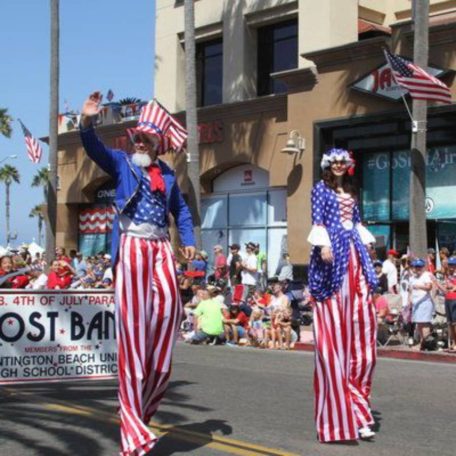 Two people on stilts in red, white, and blue costumes parade past a crowd on a sunny street, celebrating the 4th of July. American flags are visible, and a parade banner is in the background.