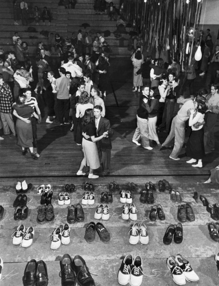 Rows of shoes line the floor in the foreground while couples dance closely in a crowded gymnasium decorated with streamers in the background. Bleachers with seated spectators are visible in the upper left.