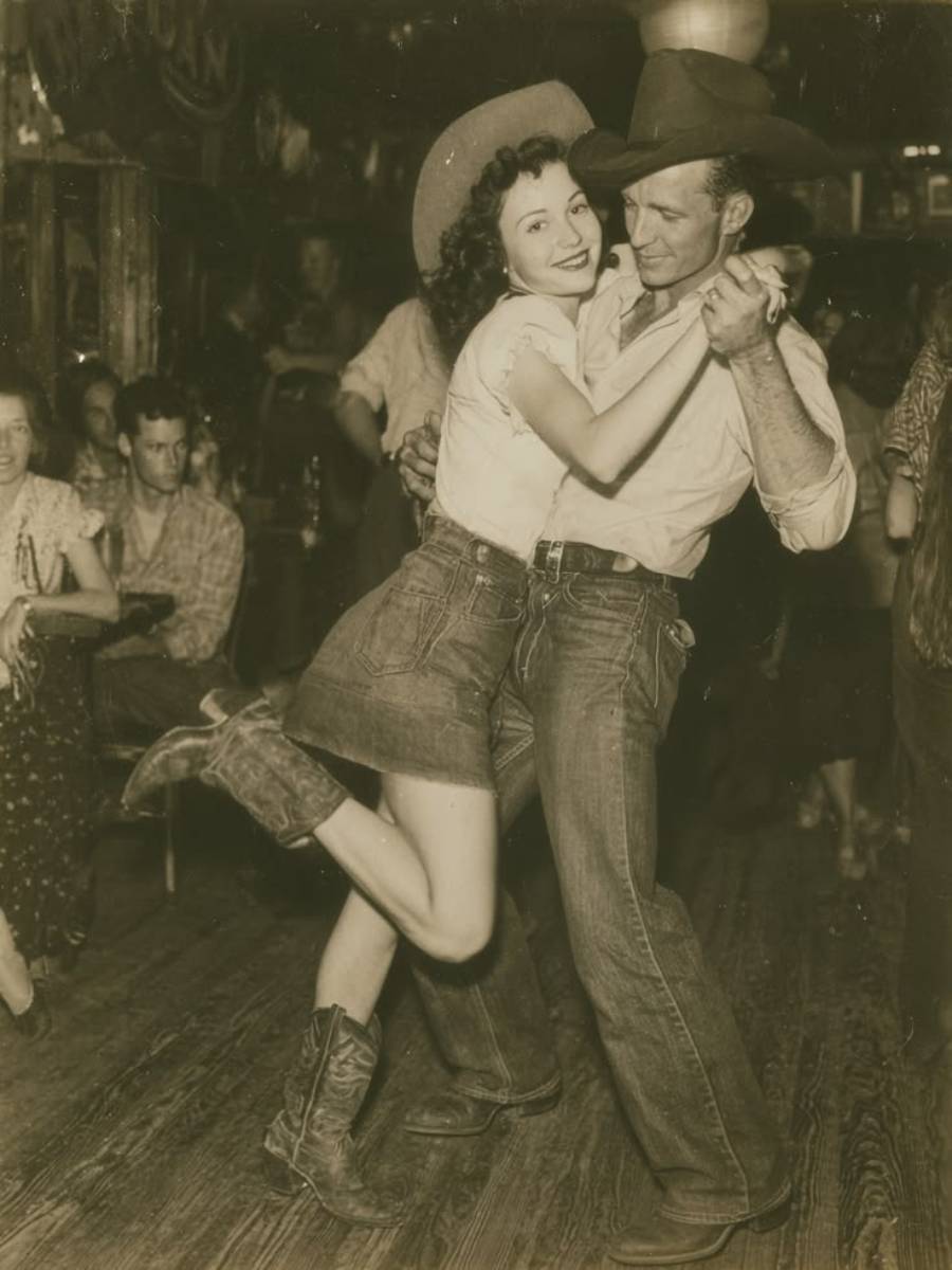 A smiling woman in a cowboy hat and boots dances with a man in jeans and a hat on a wooden floor, while people sit and watch in the background.