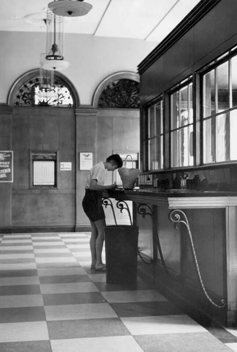 A person stands at a counter in a large, tiled room with arched windows and decorative ironwork. The scene has a vintage feel, with sunlight streaming in from above.