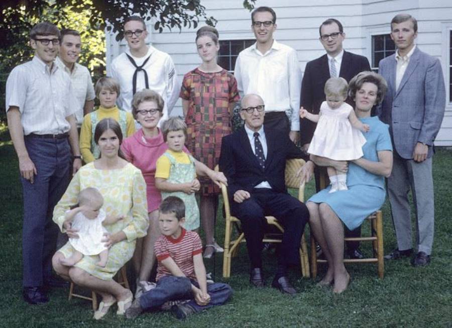 A large family poses outdoors in front of a white house, with adults standing in back and seated in front, and several children scattered among them. Everyone is dressed in vintage 1960s-style clothing.