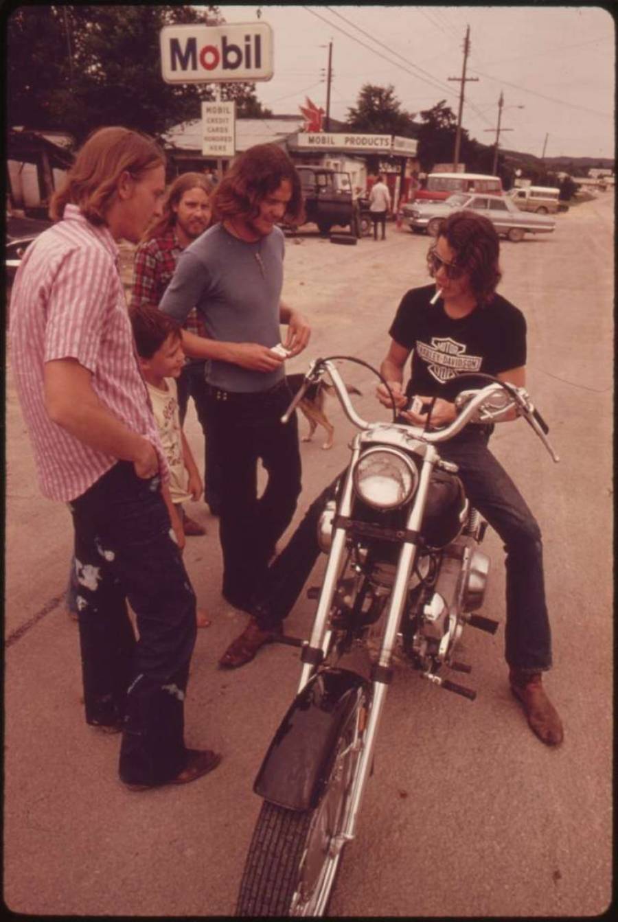 Four people stand and talk around a man sitting on a motorcycle parked on a street, with a Mobil gas station sign visible in the background.