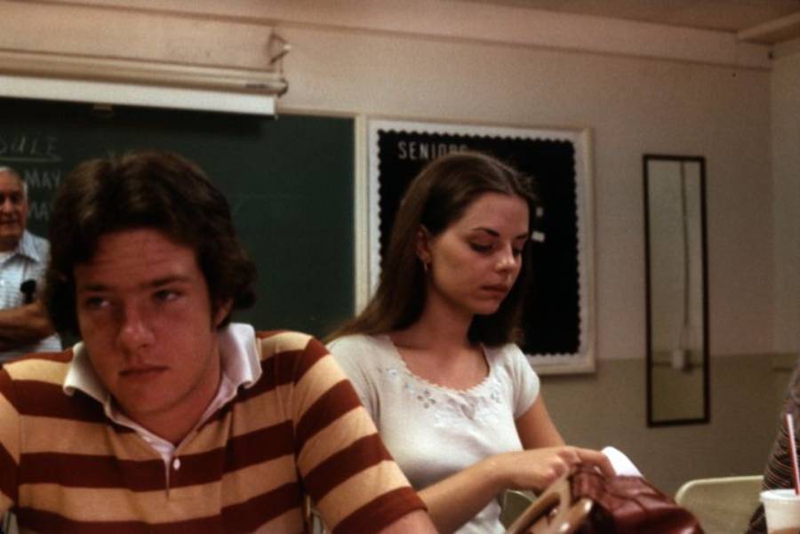Two students sit in a classroom. The boy in front wears a brown and yellow striped shirt and looks away. The girl behind him, in a white top, looks down at her desk. A chalkboard and bulletin board are visible in the background.