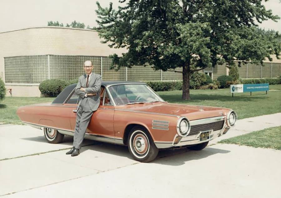 A man in a suit leans against a vintage red car parked outside a low, modern building with glass block windows and trees in the background.