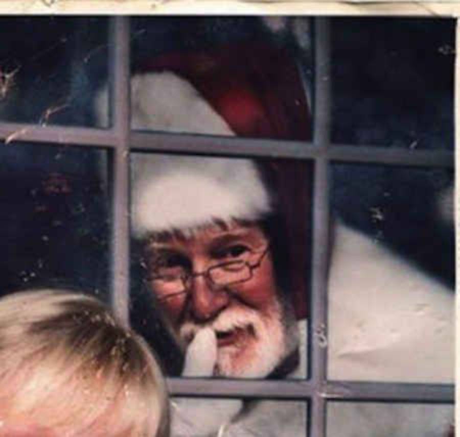 Santa Claus peeks through a window, smiling and holding a finger to his lips, signaling to be quiet. Part of a child's head is visible in the foreground.