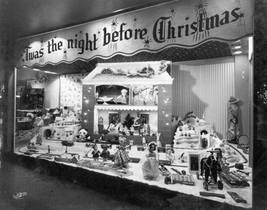 A store window displays toys and dolls beneath a banner reading "'Twas the night before Christmas." The scene is festive, with various toys arranged on shelves and snow-like decorations.