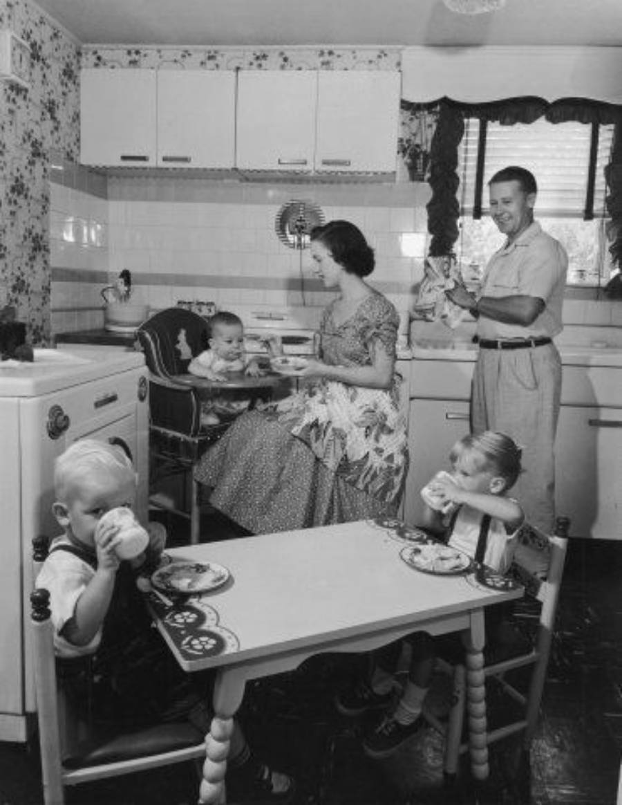 A 1950s family in a kitchen: a woman feeds a baby in a high chair, a man holds a kitten by the sink, and two young children sit at a small table drinking from cups. The room has vintage decor and patterned wallpaper.