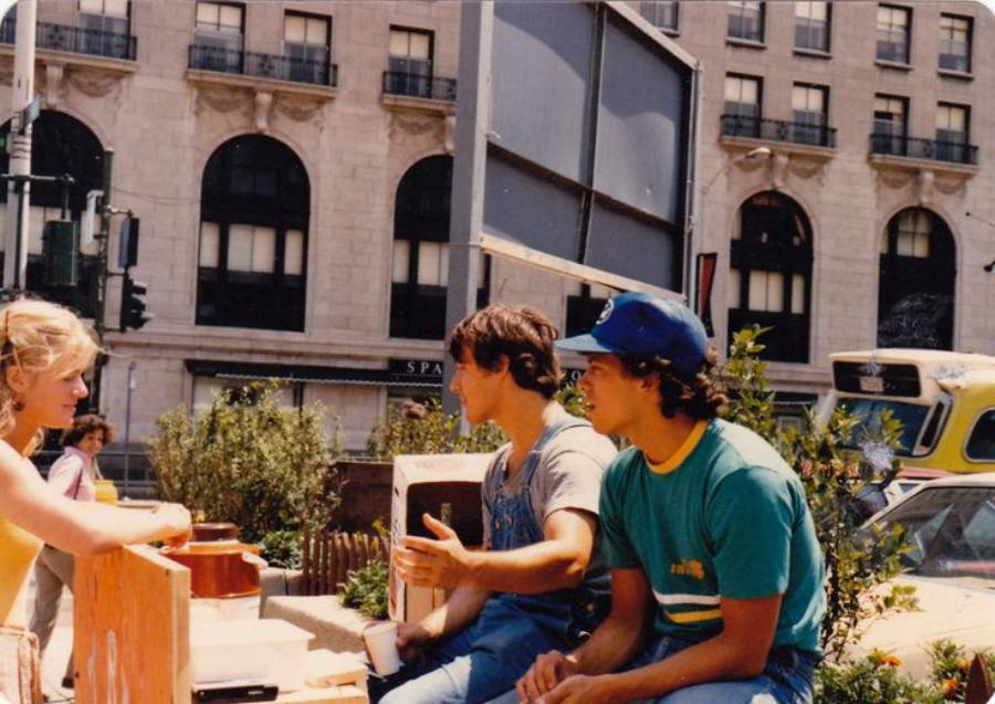 Three young adults sit and talk outdoors near a city street, with tall buildings and a large sign in the background. One woman faces two men, and a yellow vehicle is visible behind them.