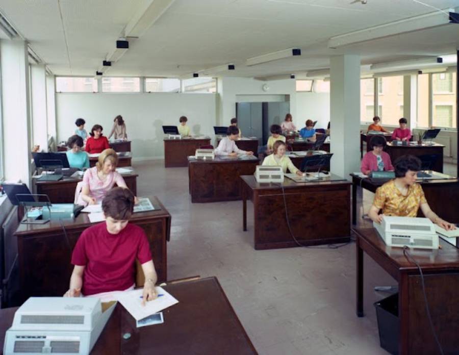 A large office room with several women and a few men sitting at individual desks, working with typewriters and documents, in a bright, mid-20th-century setting with big windows and fluorescent lighting.