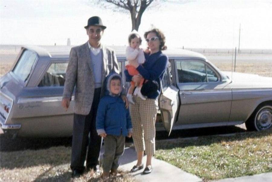 A man, woman holding a baby, and a young child stand in front of a vintage car with the door open. The scene appears to be from the 1960s, set outdoors on a sunny day with a tree and open fields in the background.