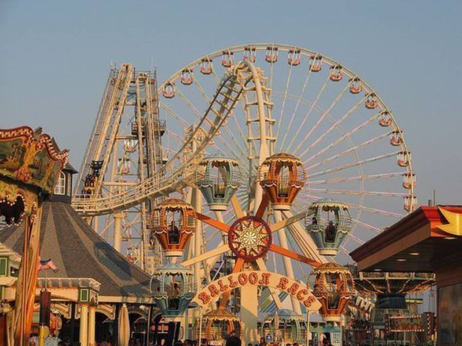 A bustling amusement park with a large Ferris wheel, roller coaster, and colorful balloon-themed ride. The scene is lively with bright attractions and structures under a clear sky.