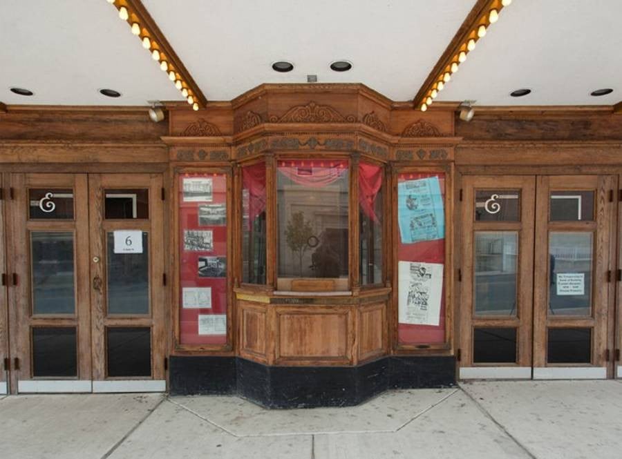 An old-fashioned theater entrance with a central wooden ticket booth, glass display cases with vintage posters and photos on both sides, and four doors; the marquee is lined with small lights.