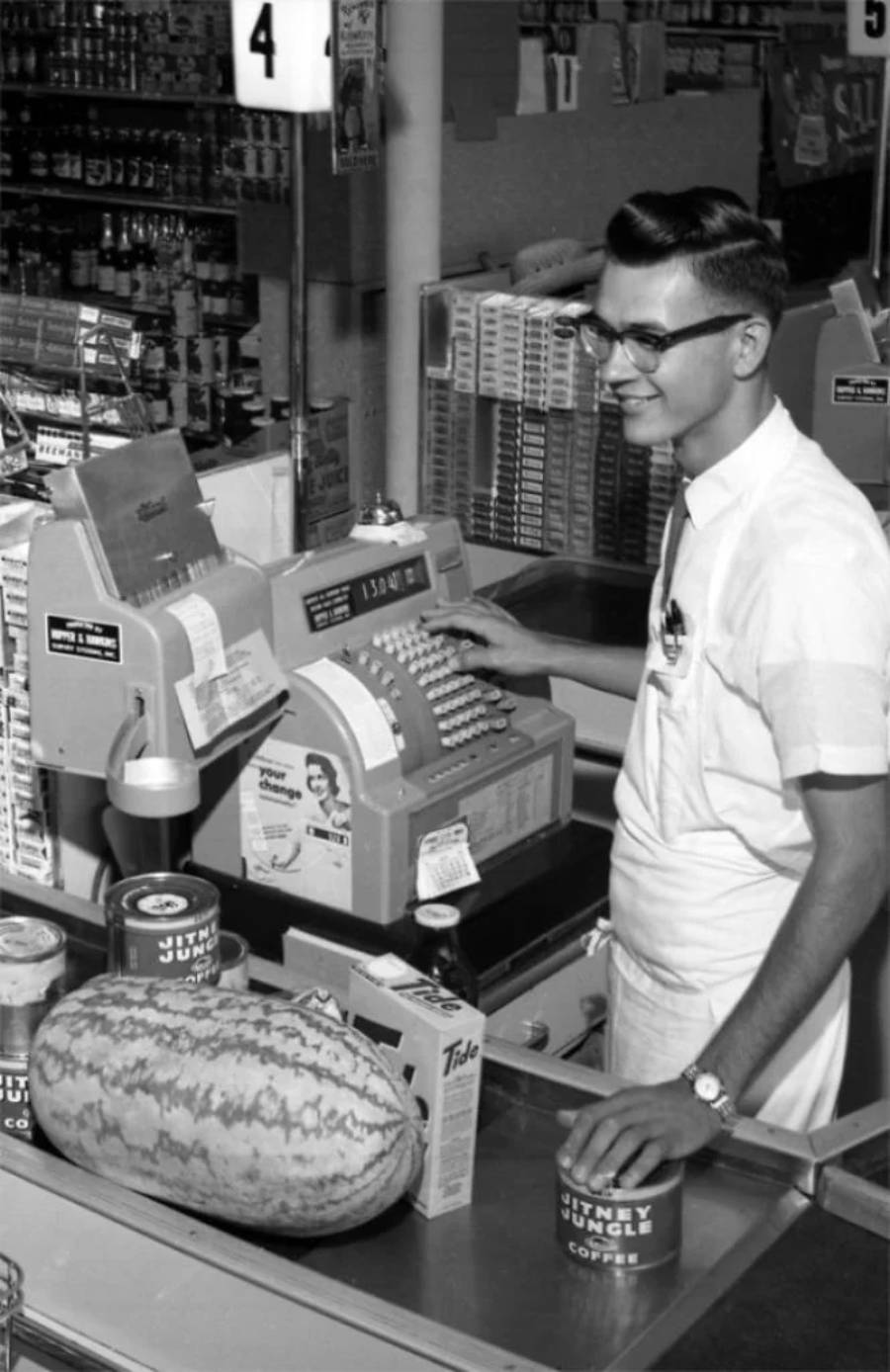 A young male cashier in glasses and a white uniform rings up groceries, including a watermelon and canned coffee, at an old-fashioned cash register in a vintage grocery store.