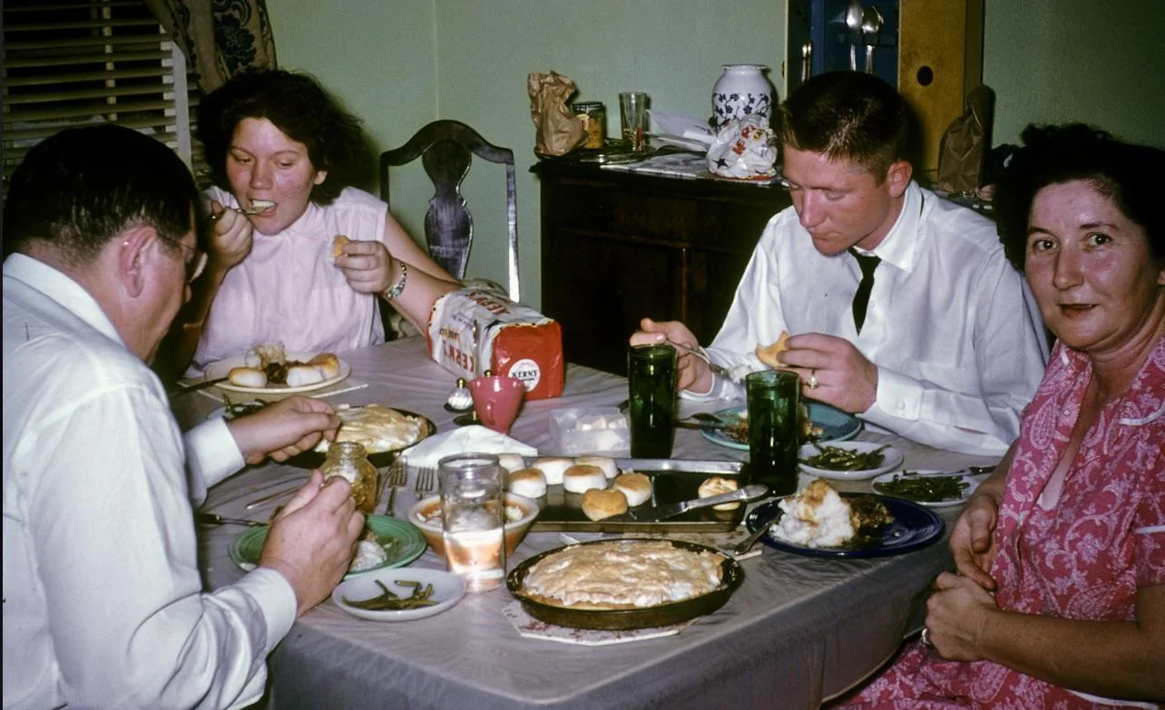 Four adults sit around a dining table eating a meal together. The table is set with pies, bread rolls, green beans, drinks, and various dishes. One woman looks at the camera while others are eating.