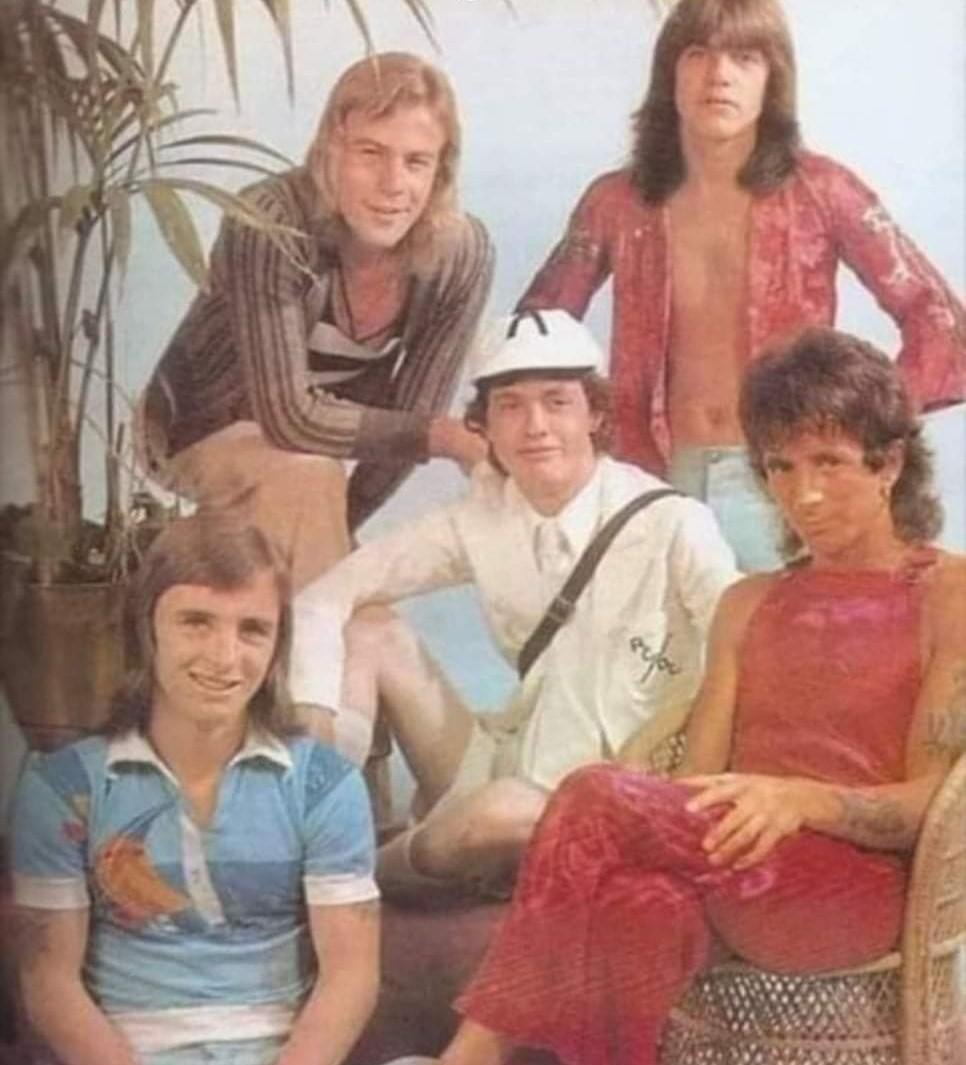 Five men pose together indoors by a tall plant; three are seated and two stand behind. They wear colorful, casual 1970s clothing, with one in a white hat and outfit, and others in patterned shirts and pants.