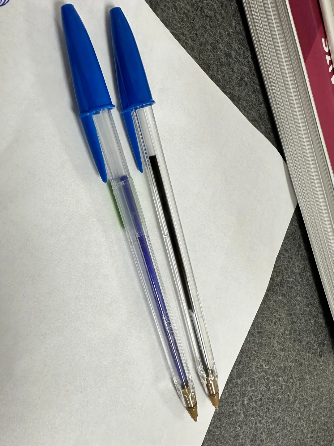Two clear ballpoint pens with blue caps rest on a white sheet of paper, next to the edge of a thick book with a reddish cover, all placed on a gray surface.