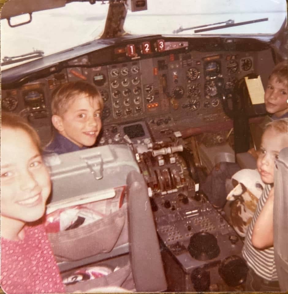 Three smiling children sit in an airplane cockpit, surrounded by controls and buttons. One child holds a stuffed animal. The scene appears to be from the past, suggested by the photo’s vintage quality.