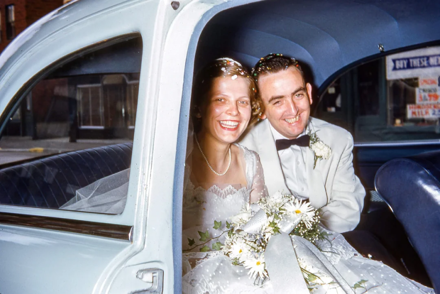 A smiling bride and groom in wedding attire sit in the backseat of a vintage car, holding a bouquet of white flowers.