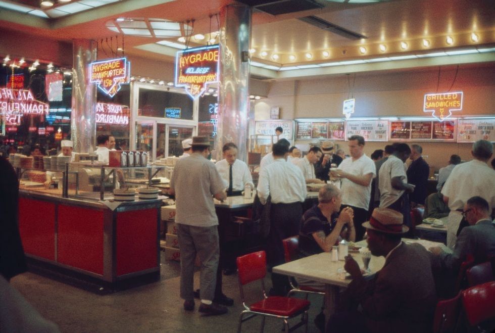 A busy diner with neon signs, people ordering at the counter, and others sitting at red chairs and tables, eating and talking. The scene has a retro, mid-20th-century atmosphere.