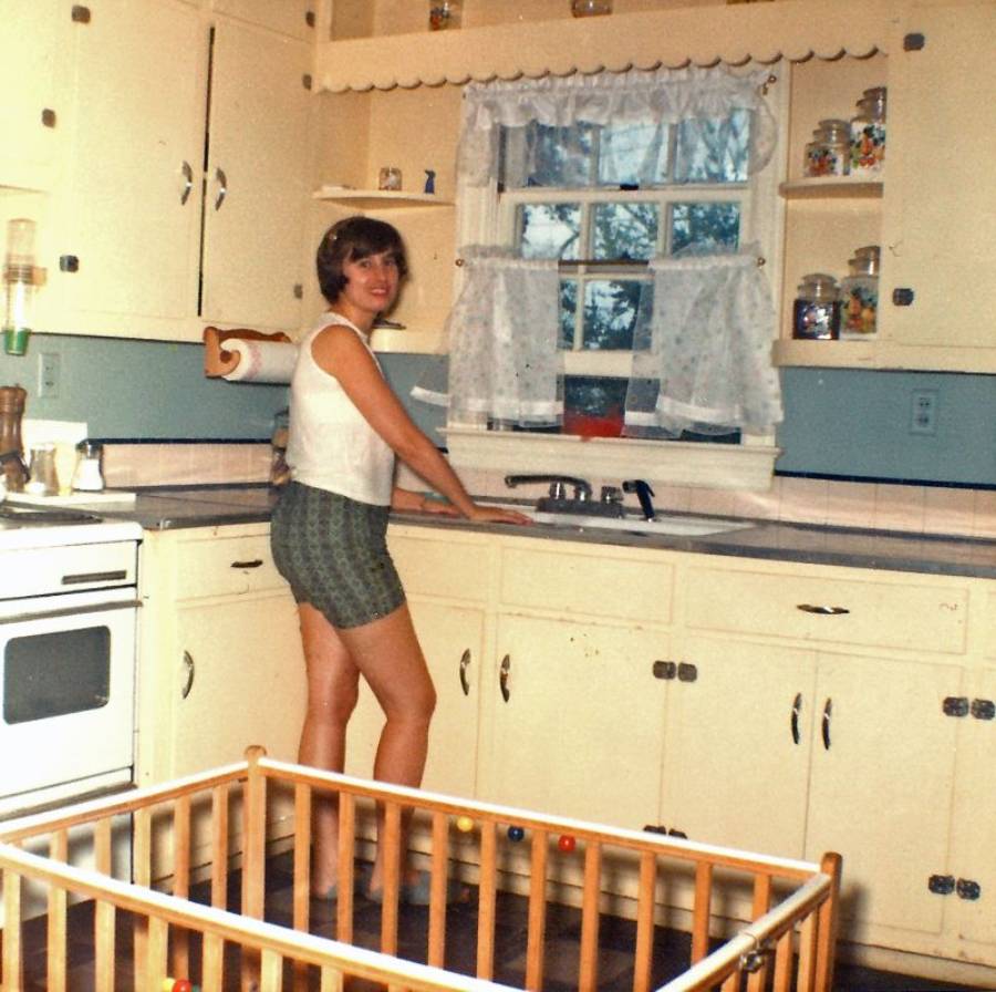 A woman stands at a yellow kitchen sink, smiling, with white cabinets and a window with lace curtains behind her. A baby playpen is visible in the foreground. The setting appears vintage.