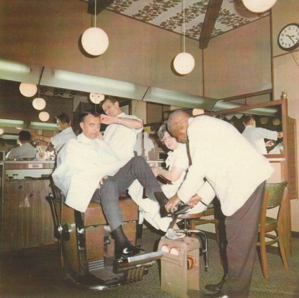 A man gets his hair cut and shoes shined at the same time in a vintage barbershop, with two barbers working and mirrors reflecting the scene.
