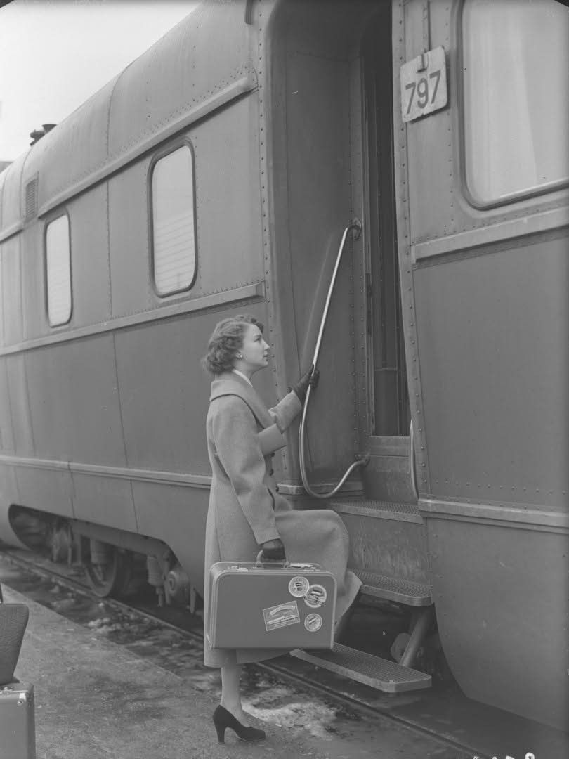 A woman in a coat and heels holding a suitcase steps onto a train. The train car has the number 797 and travel stickers on the suitcase. The scene appears to be from a past era.