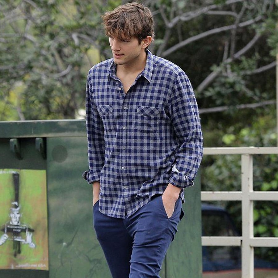 A man with tousled brown hair wears a blue plaid shirt and navy pants, walking outdoors with his hands in his pockets. Trees and a green dumpster are in the background.