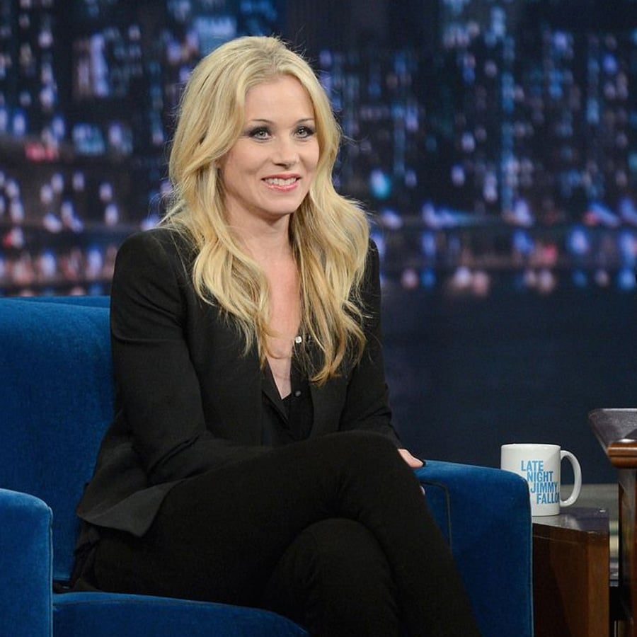 A woman with long blonde hair, dressed in black, sits on a blue chair during a talk show. A mug labeled "Late Night with Jimmy Fallon" is on the table beside her.