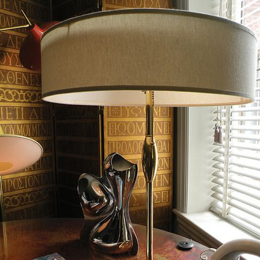 A brass floor lamp with a beige shade stands beside a window with blinds. Behind it, a wall is decorated with golden Greek letters. A silver abstract sculpture and a pink lamp are on a polished round wood table.