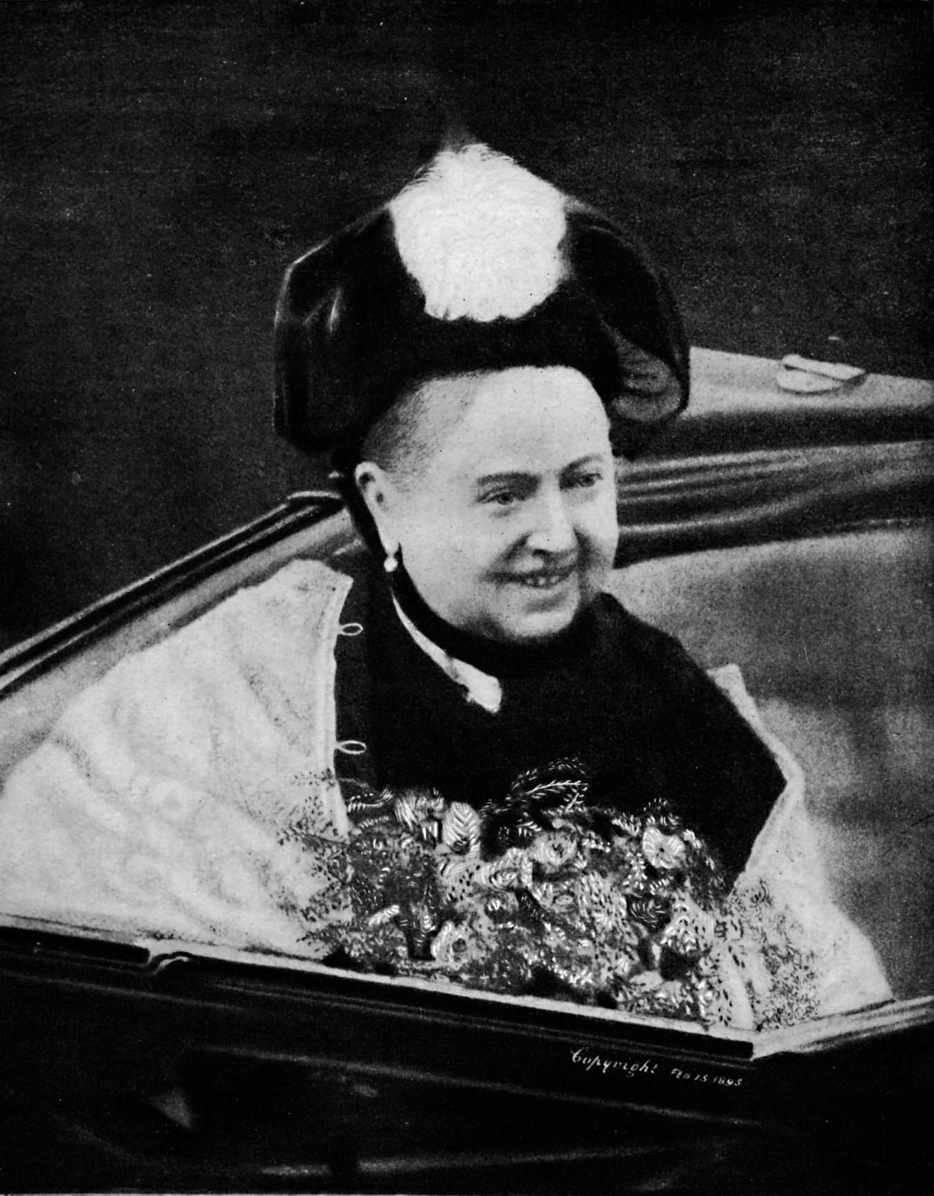 An older woman in ceremonial dress with a feathered hat sits in an open carriage, smiling and holding a large bouquet of flowers.