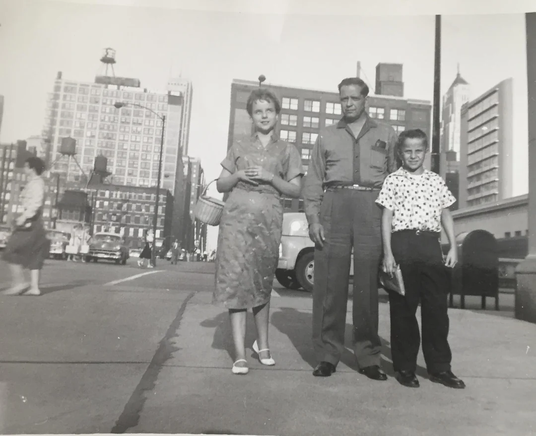 A vintage black and white photo of a woman, man, and boy standing on a city sidewalk with tall buildings, cars, and pedestrians in the background. The boy holds a book while the adults stand close together.