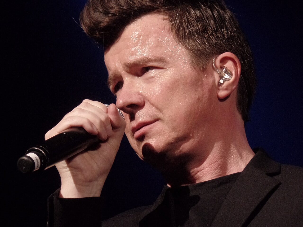 A man with short brown hair sings into a microphone on stage, wearing a black outfit and an in-ear monitor, with sweat visible on his face and a dark background behind him.