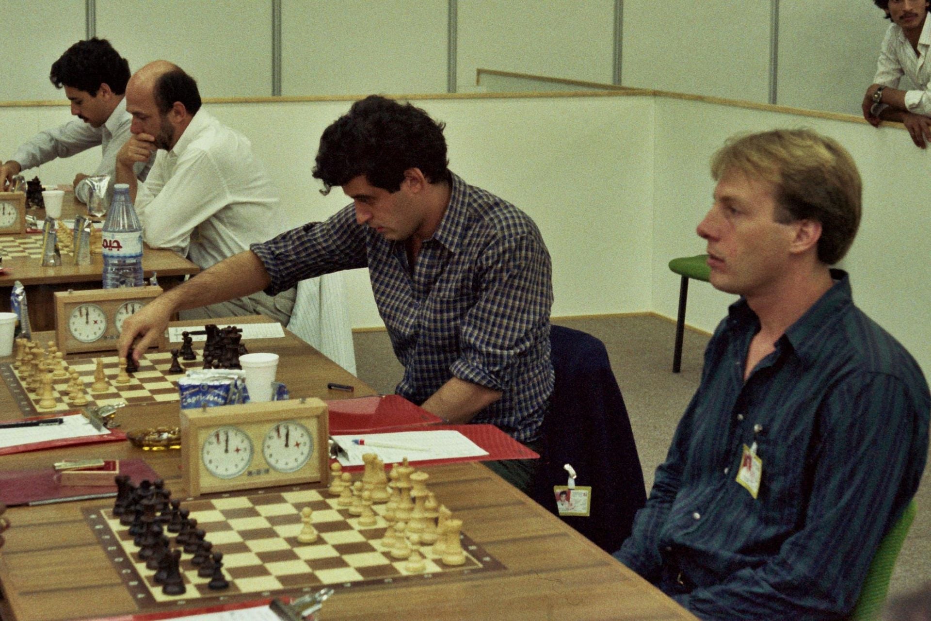 Four men sit at two chessboards, concentrating. Two chess clocks are visible. One man moves a piece while another sits across from him. Two more men observe or play nearby. The setting appears to be a chess tournament.
