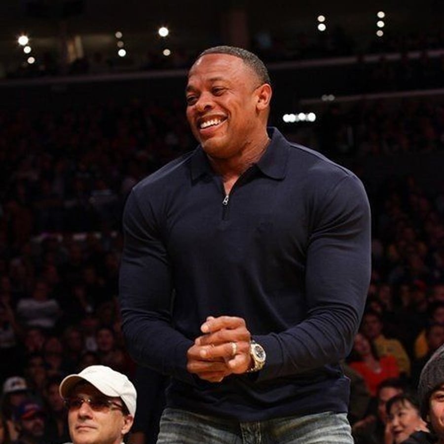 A man in a navy long-sleeve shirt stands and smiles in a crowded indoor arena, with people seated around him.